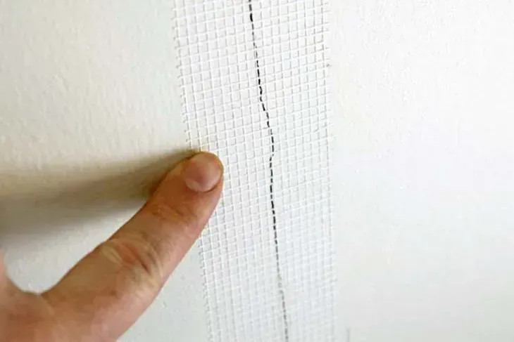 Finger pointing at a wall crack covered with white mesh tape.