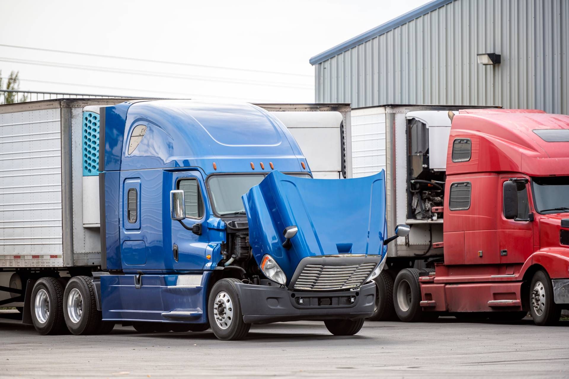 Blue semi-truck with open hood, next to a red truck and white trailers, parked outside a warehouse.