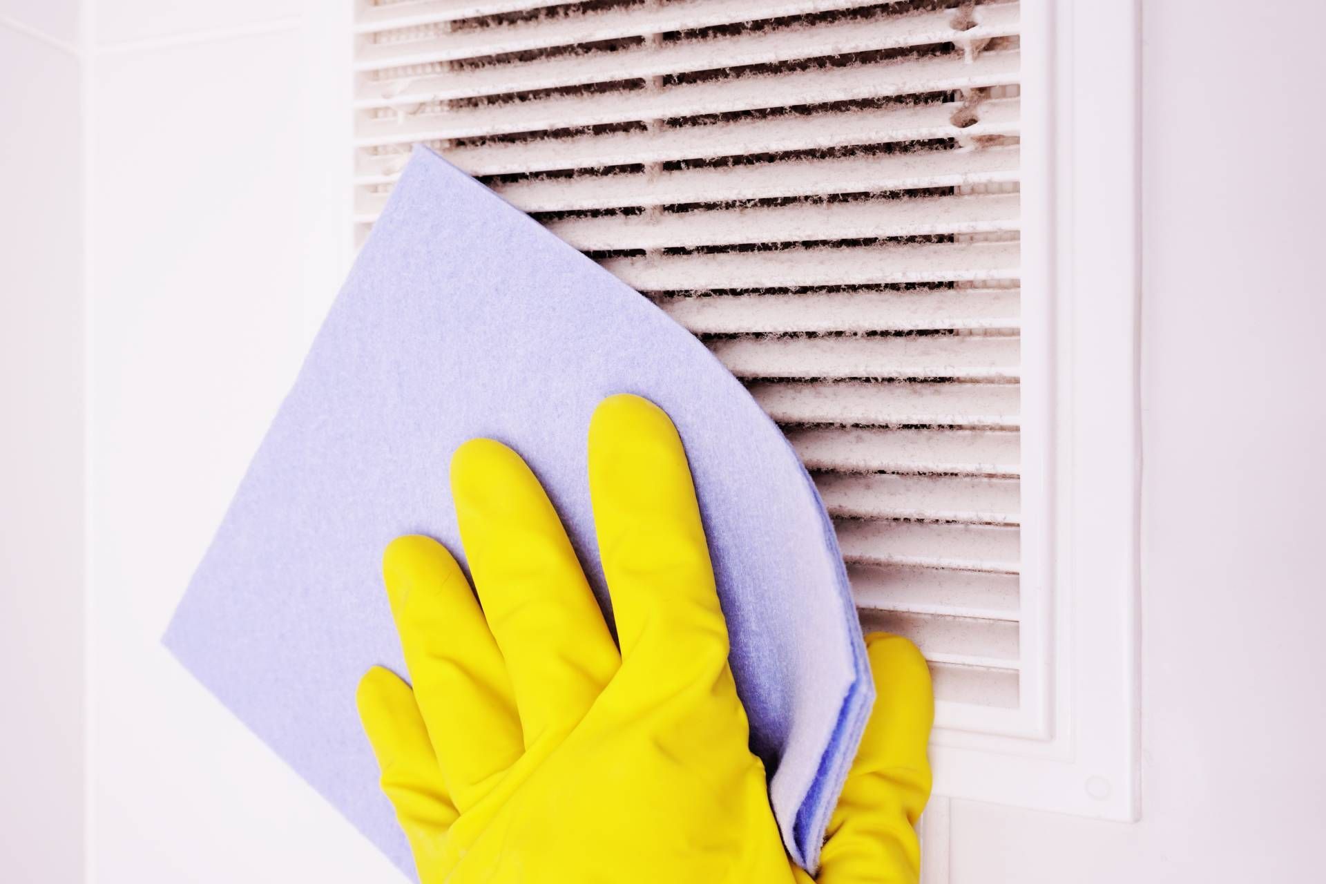 A person wearing yellow gloves cleaning a dusty air vent with a blue cloth.