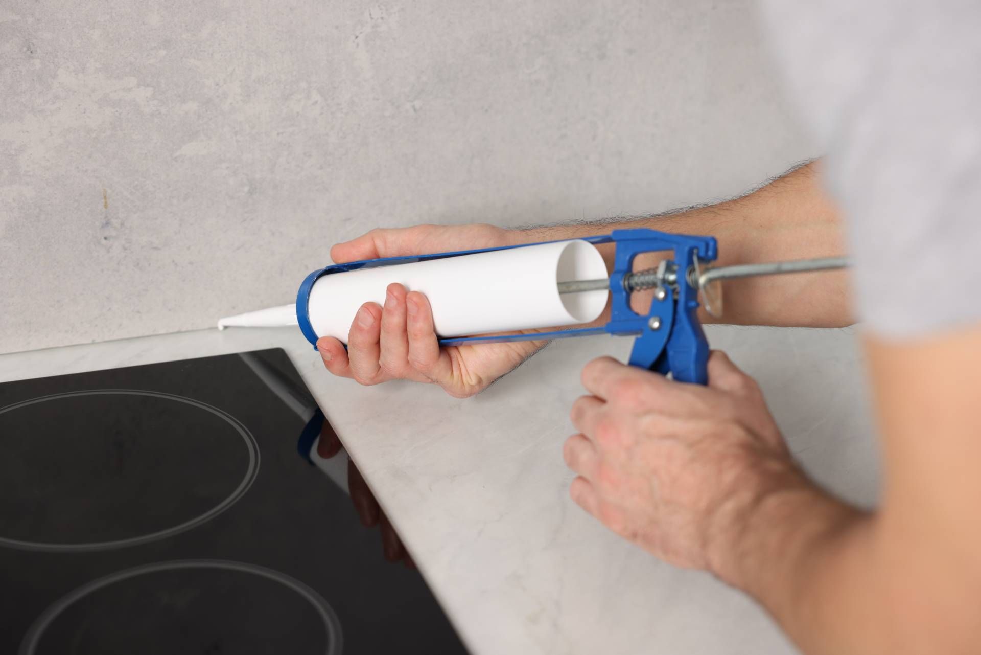 Person applying caulk with a blue caulking gun to a countertop near a stovetop.
