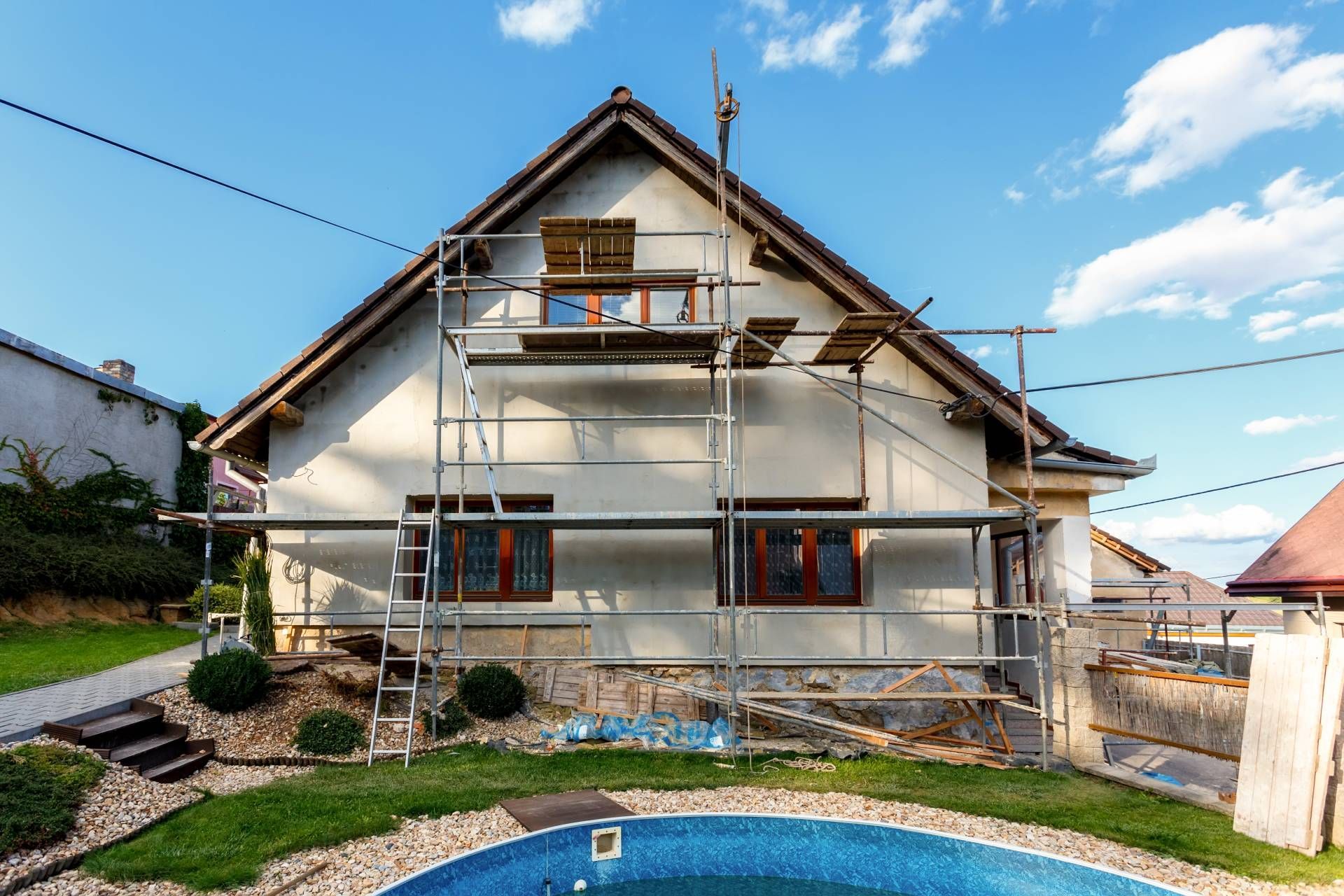 House exterior under renovation; scaffolding, pool in foreground, blue sky.