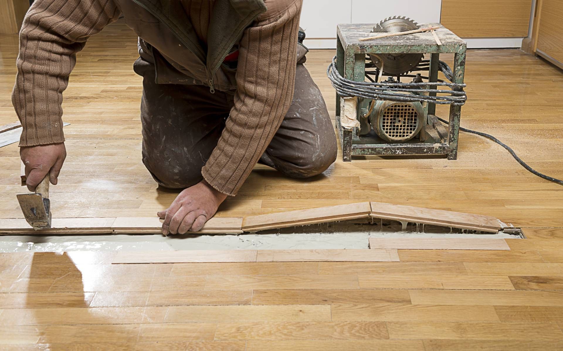 Person applying adhesive to wood flooring with a trowel, a saw in the background.