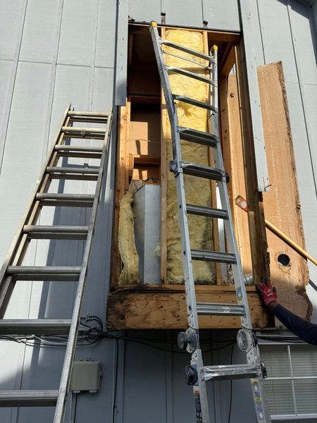 Ladder against house wall, opening with insulation, person's hand removing wood.