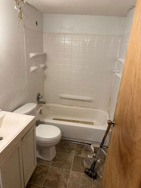 Bathroom with white tub/shower, toilet, and sink, brown floor, door ajar.