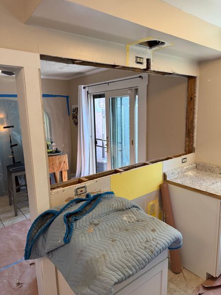 Kitchen remodel: wall opening to a room with a patio door. Electrical wiring visible. Protective blanket on cabinet.