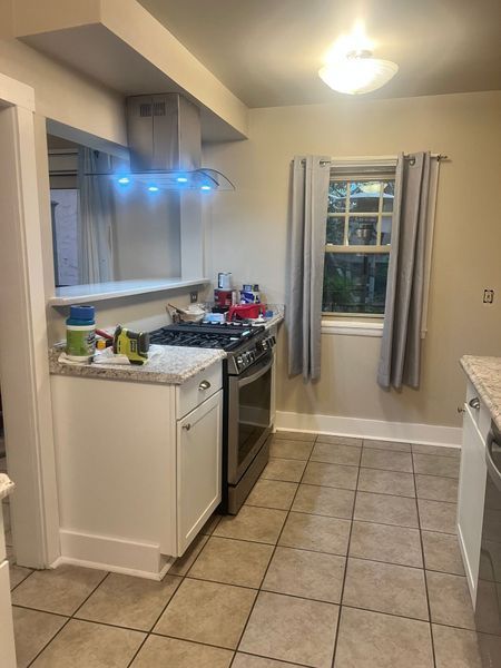 Kitchen with stove, cabinets, window with gray curtains. Beige floor tiles.