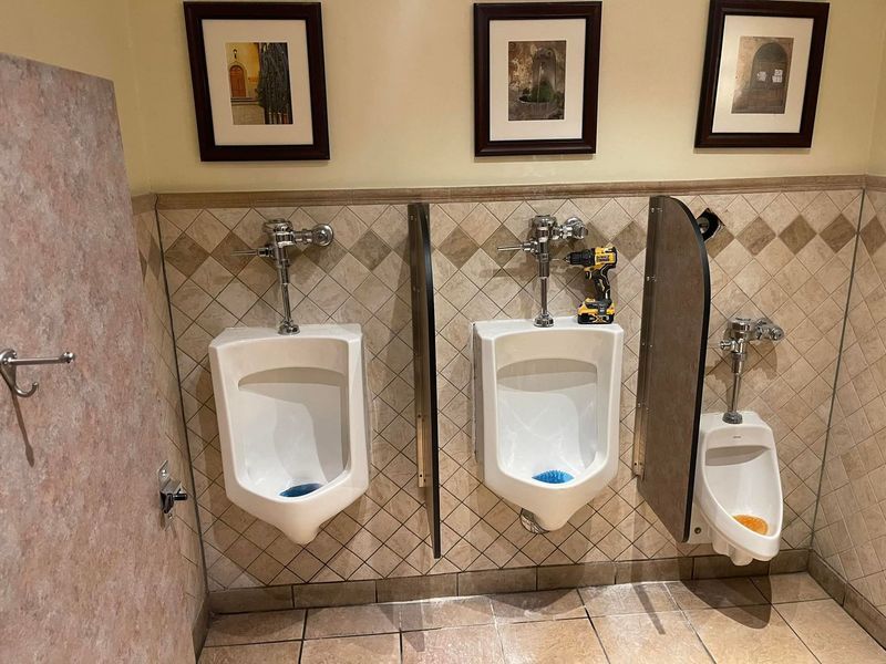 Public restroom with three urinals. The middle urinal has a drill resting on it. Beige walls and patterned tile.