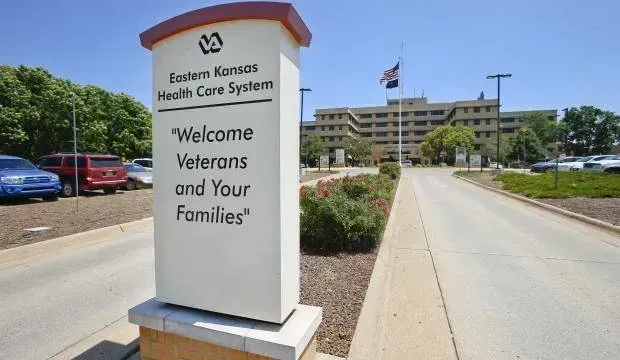 Sign for Eastern Kansas Health Care System welcomes veterans and families; building and parking lot in background.