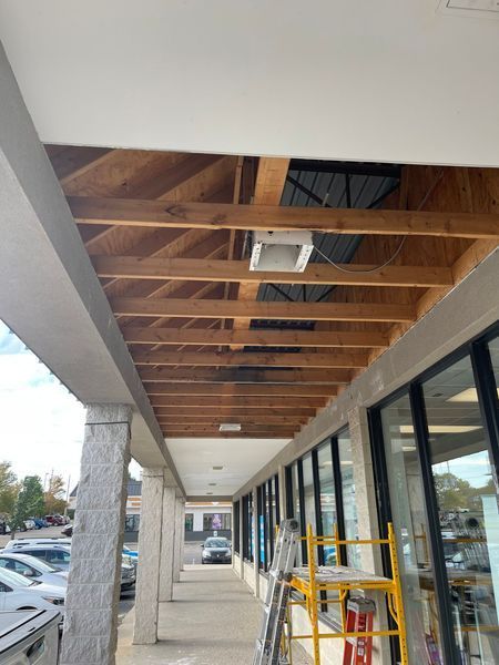 Exterior view of a building under construction, revealing ceiling beams. A walkway with storefronts is visible.