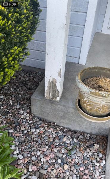White porch pillar with peeling paint, next to a potted plant and gravel.