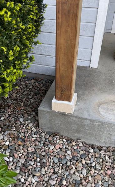 Wooden post stained brown, attached to a concrete base, with gravel and greenery surrounding it.