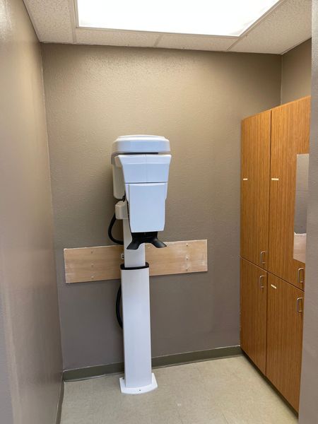 Dental x-ray machine in a beige-walled room, next to wooden cabinets.