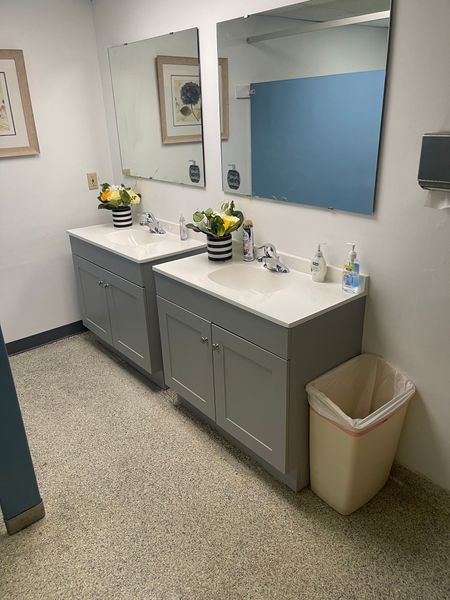 Two gray bathroom vanities with mirrors, flowers, and a wastebasket.