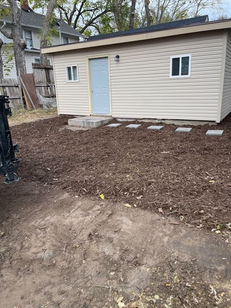 A tan shed with a light blue door and two windows in a yard covered in mulch.