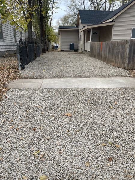 Gravel driveway leading to a house with a wooden fence and gray siding. Cloudy sky.