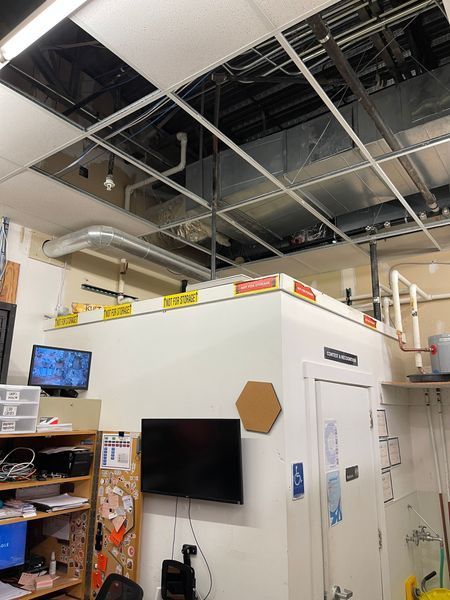 Ceiling with missing tiles exposing ducts and pipes. Below, a white room with a door, TV screens, and shelves.