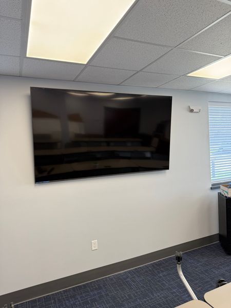 Large TV mounted on a white wall in a room with a blue carpet and a ceiling with fluorescent lights.