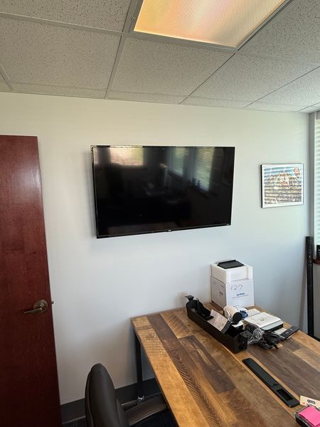 Large, flat-screen TV mounted on a white office wall above a wooden desk; door on left, window on right.