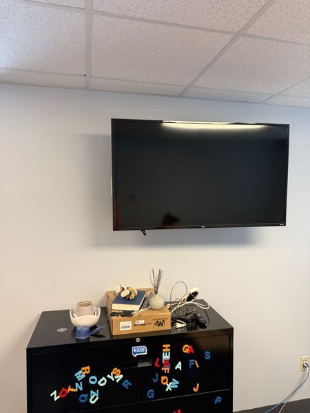 A television mounted on a white wall above a black dresser decorated with colorful letters and objects.