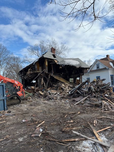 Burned-out house in rubble; a skid steer and a blue dumpster are visible. Cloudy sky.