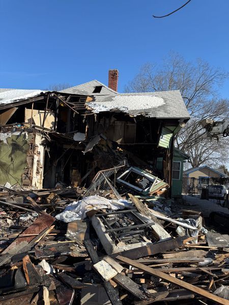 A house severely damaged by fire, with charred wood and debris scattered under a blue sky.