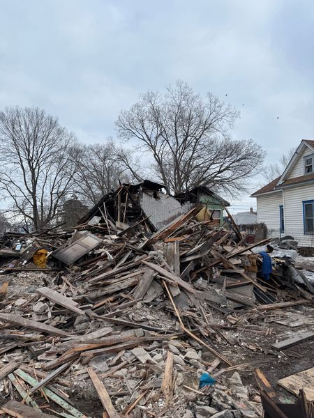 Debris pile from a demolished building. Gray wood and rubble with visible tree branches against a cloudy sky.