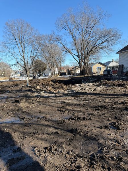 A muddy, bare lot with several leafless trees and houses in the background under a blue sky.