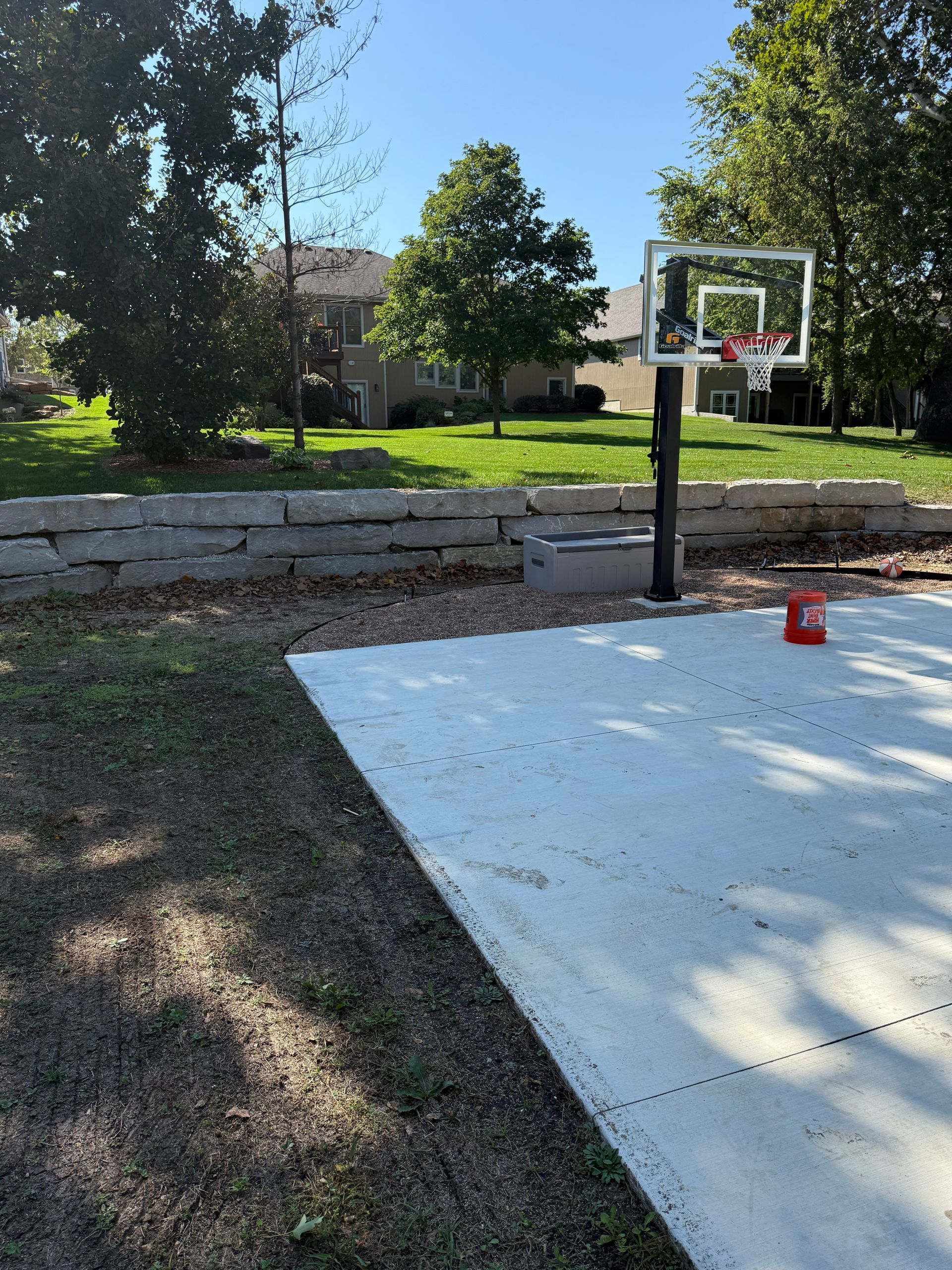 Basketball hoop on a concrete court in a backyard. Trees, grass, and a retaining wall are in the background.