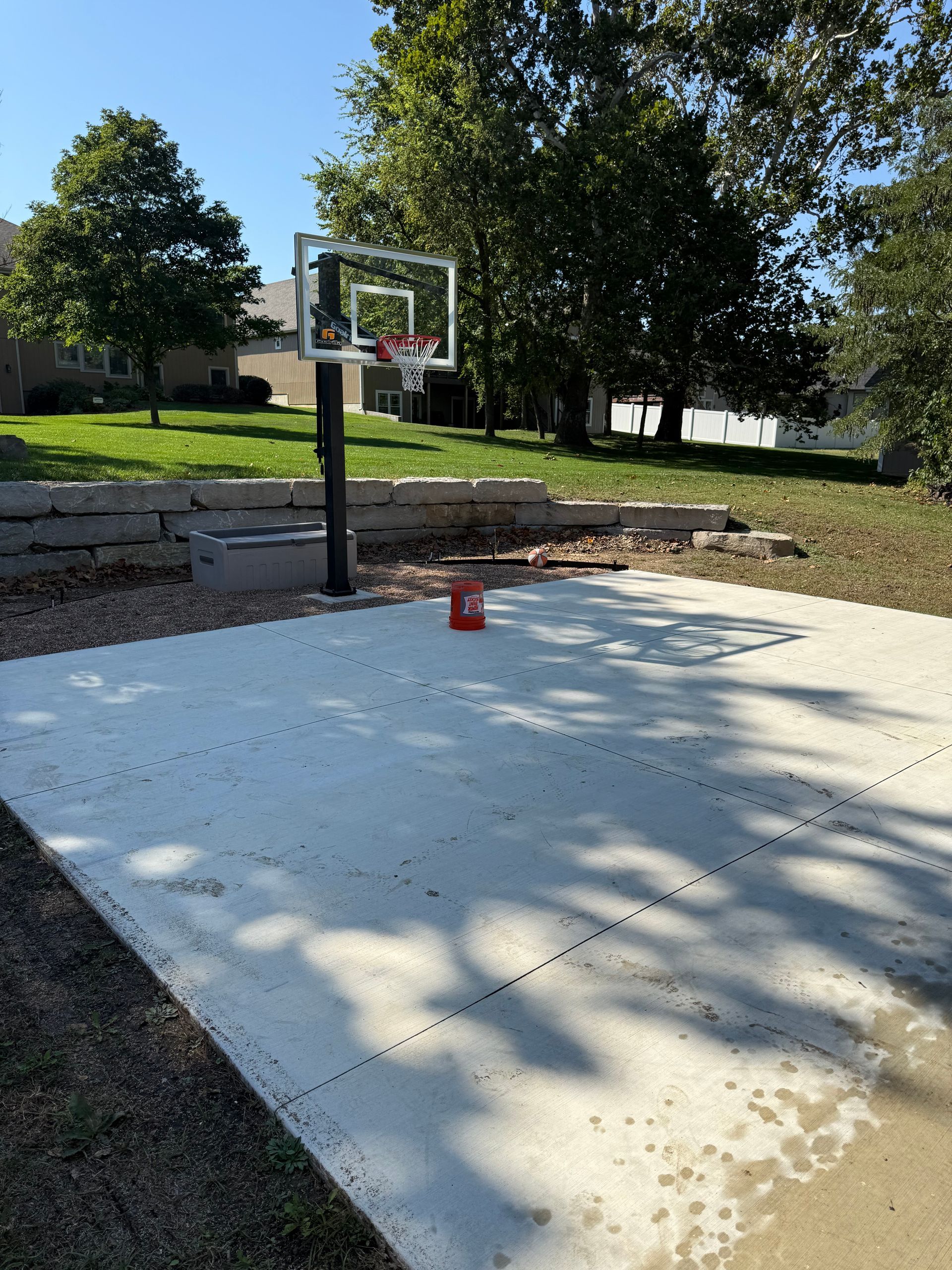 Basketball hoop on a concrete court in a backyard. Green grass and trees in the background.