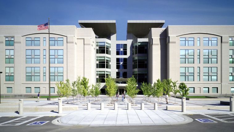 Modern building with a central glass section, stone facade, US flag, and trees.