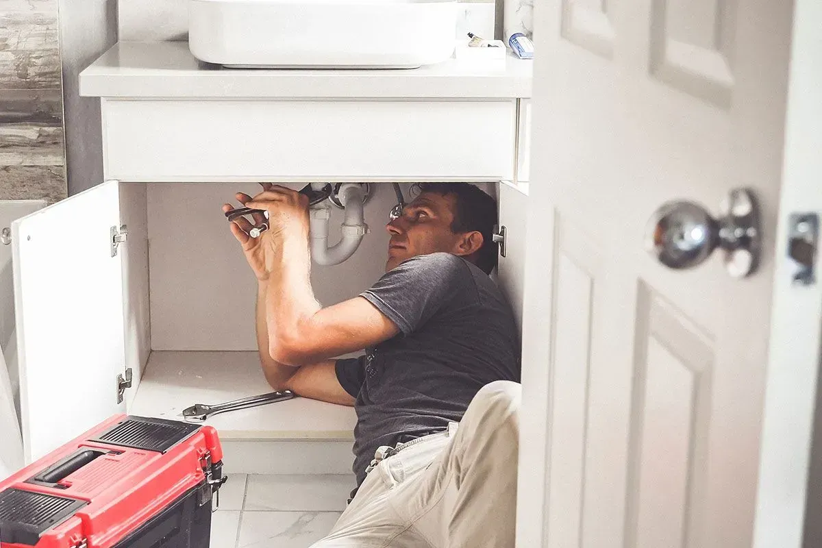 A man in a bathroom repairs pipes under a sink with tools.
