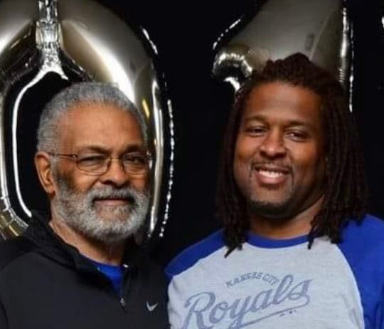 Two men smiling, one with gray beard and glasses, the other with dreadlocks, in front of silver balloons.