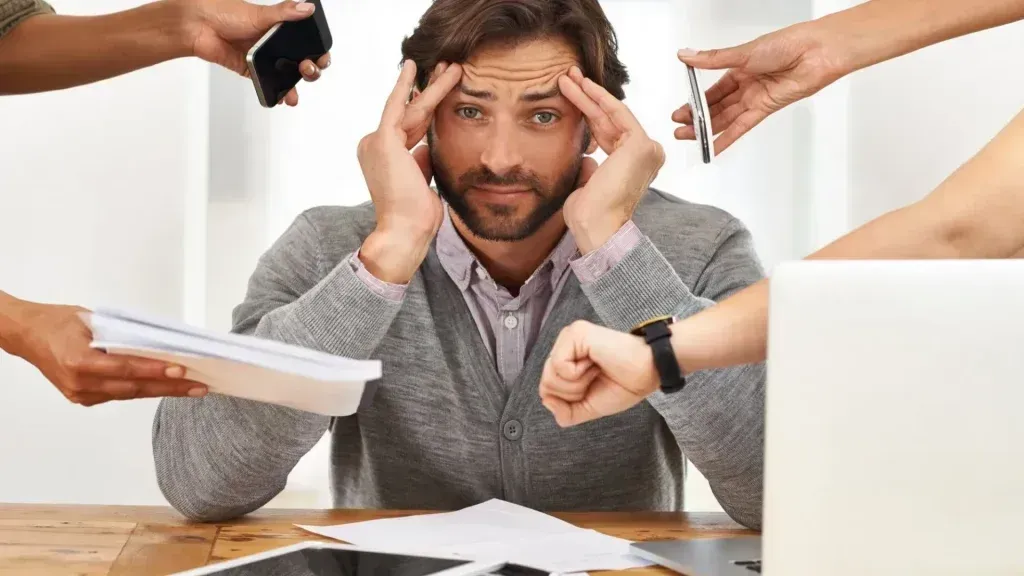 Man stressed, surrounded by hands holding documents, phones, and a watch.