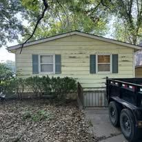 Yellow house with peeling paint, a trailer, and overgrown yard.