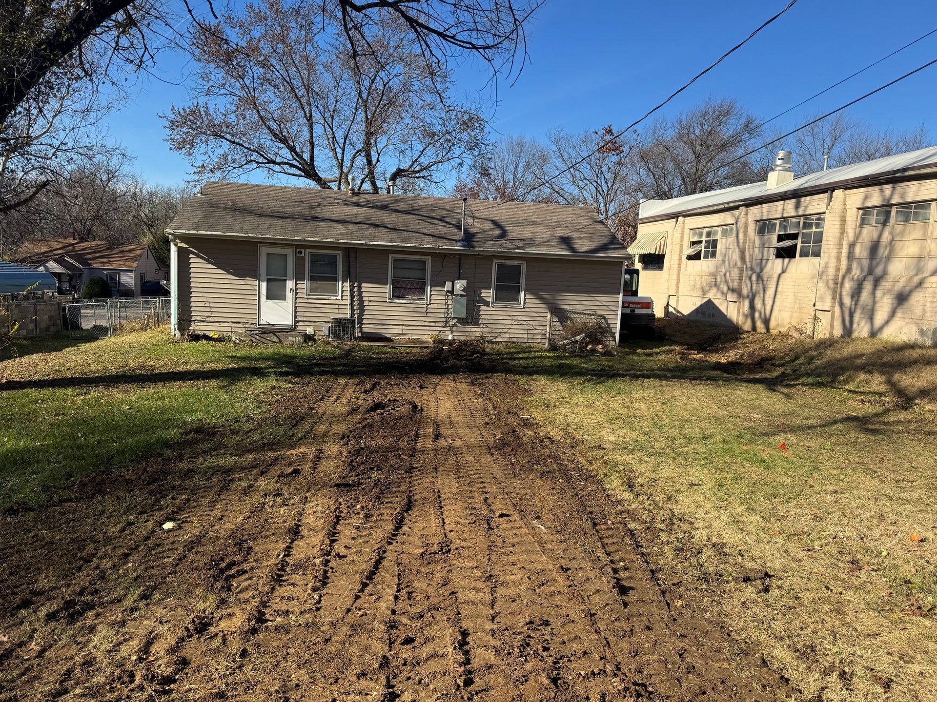 Dirt path leads to a beige house and building under a blue sky. Bare trees and grass surround.
