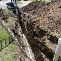 Trench dug for underground utility lines. Backhoe nearby. Pipes visible in trench.