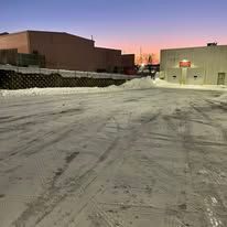 Snow-covered parking lot at dusk with industrial buildings in the background.