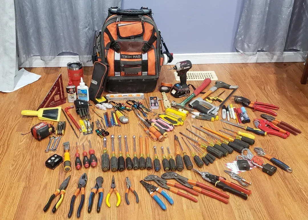 An assortment of electrician tools laid out on a wood floor, with a tool backpack in the background.
