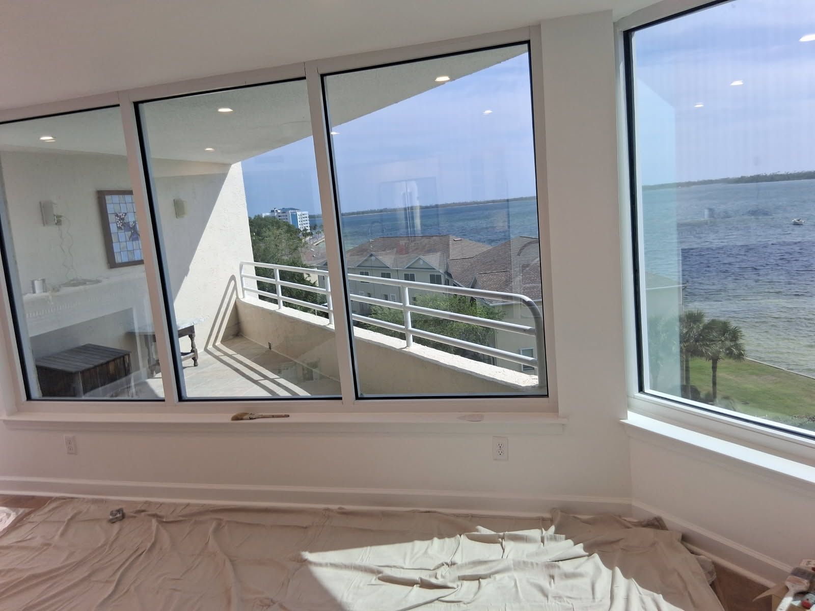 Interior view of a room with large windows overlooking water and a balcony. White walls and floors, sunlight.