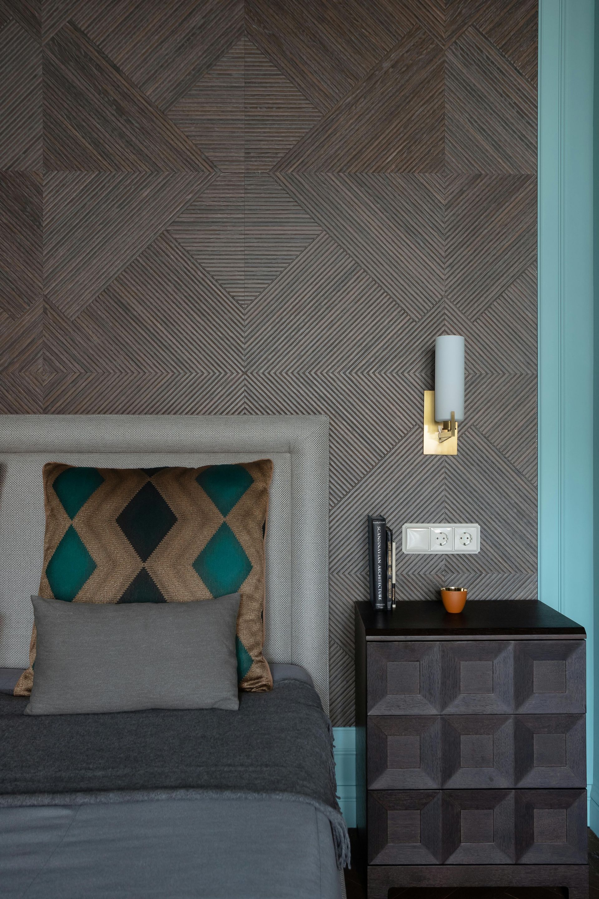 Bedroom with patterned gray wall, bed with teal and gray pillows, nightstand, and sconce.