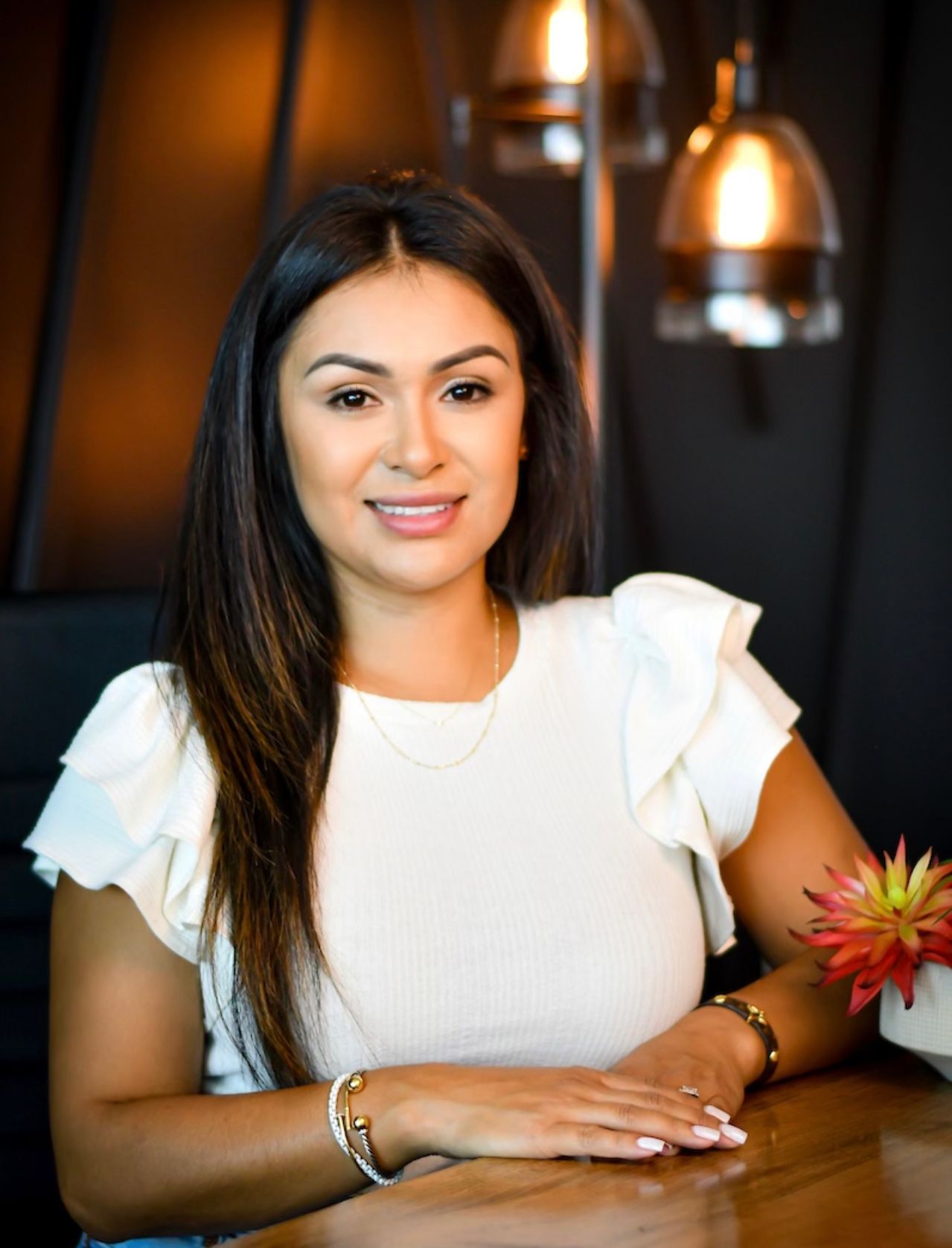 Woman with long dark hair, smiling, seated at a wooden table in front of a dark background with hanging lights.