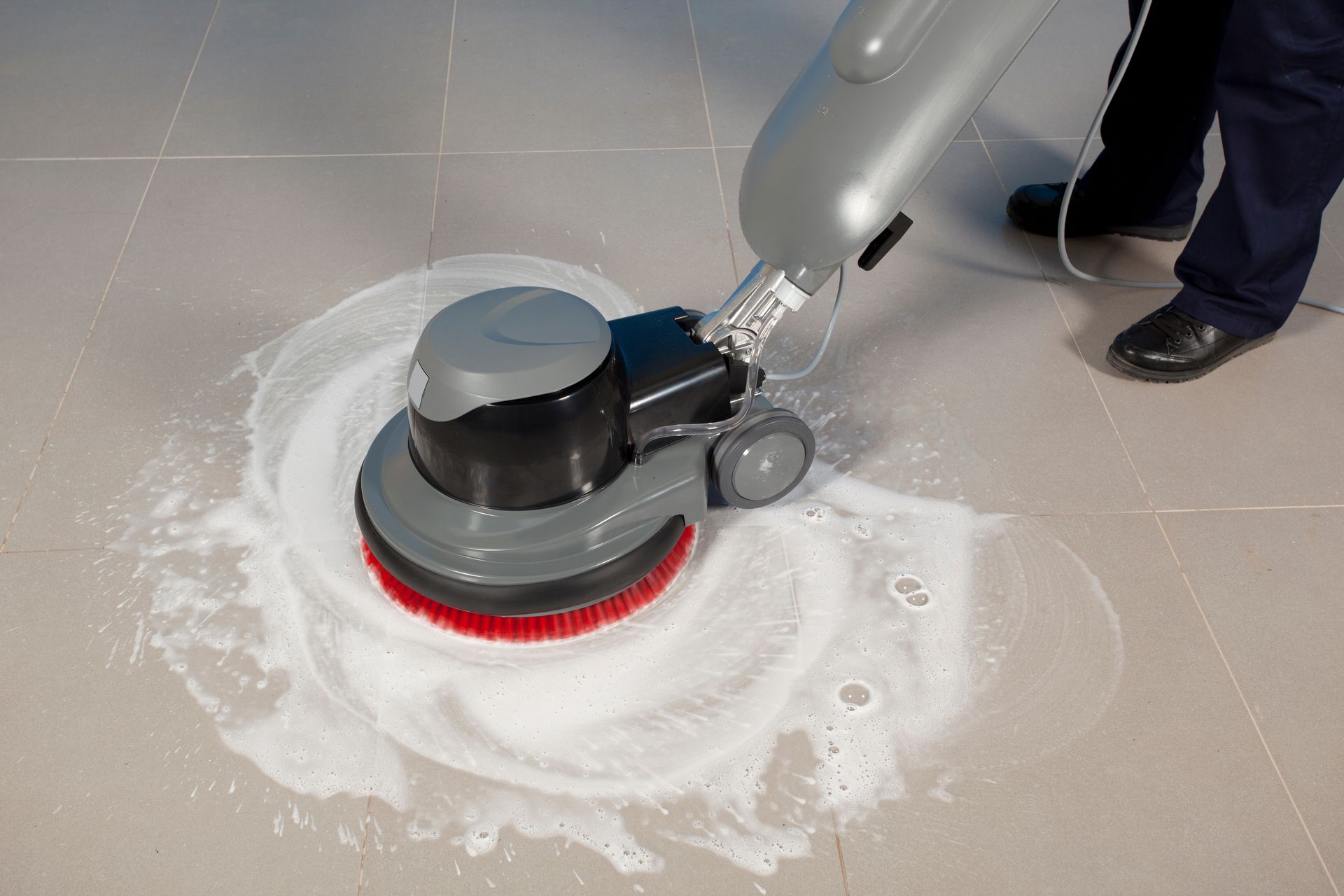 Floor scrubber cleaning a tiled floor, leaving white foam around the rotating brush. Floor scrubber cleaning a tiled floor, spreading white foam around the machine