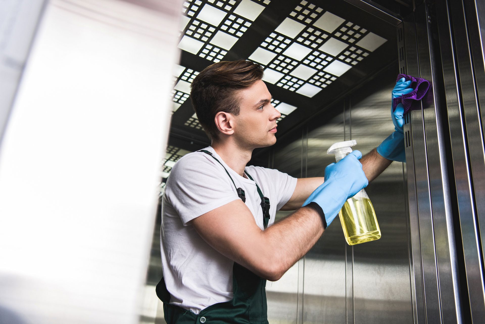 Worker cleaning a stainless steel surface with blue gloves and spray bottle in a bright industrial kitchen