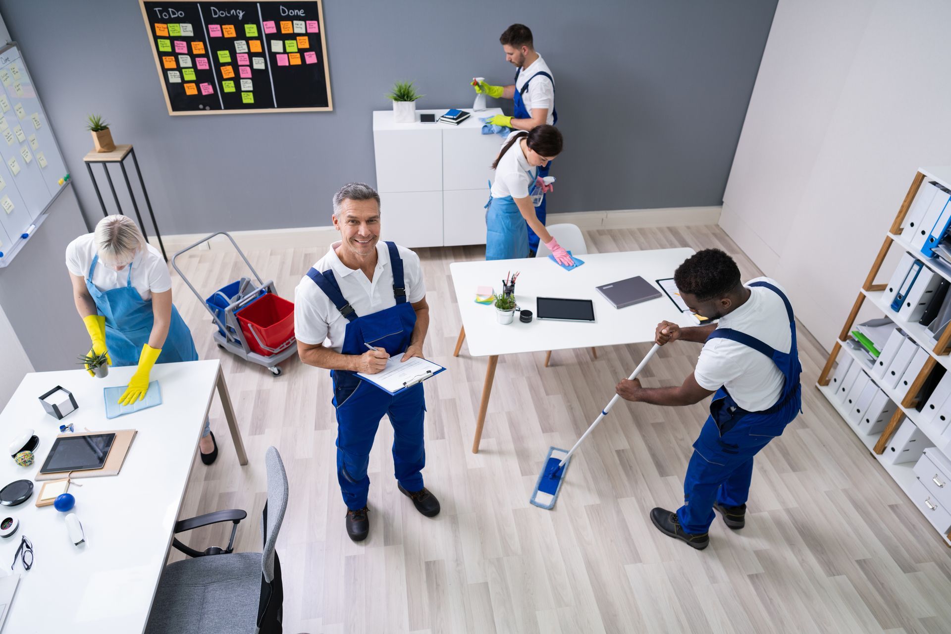 People cleaning a modern office with mops, buckets, and supplies around desks and shelves.