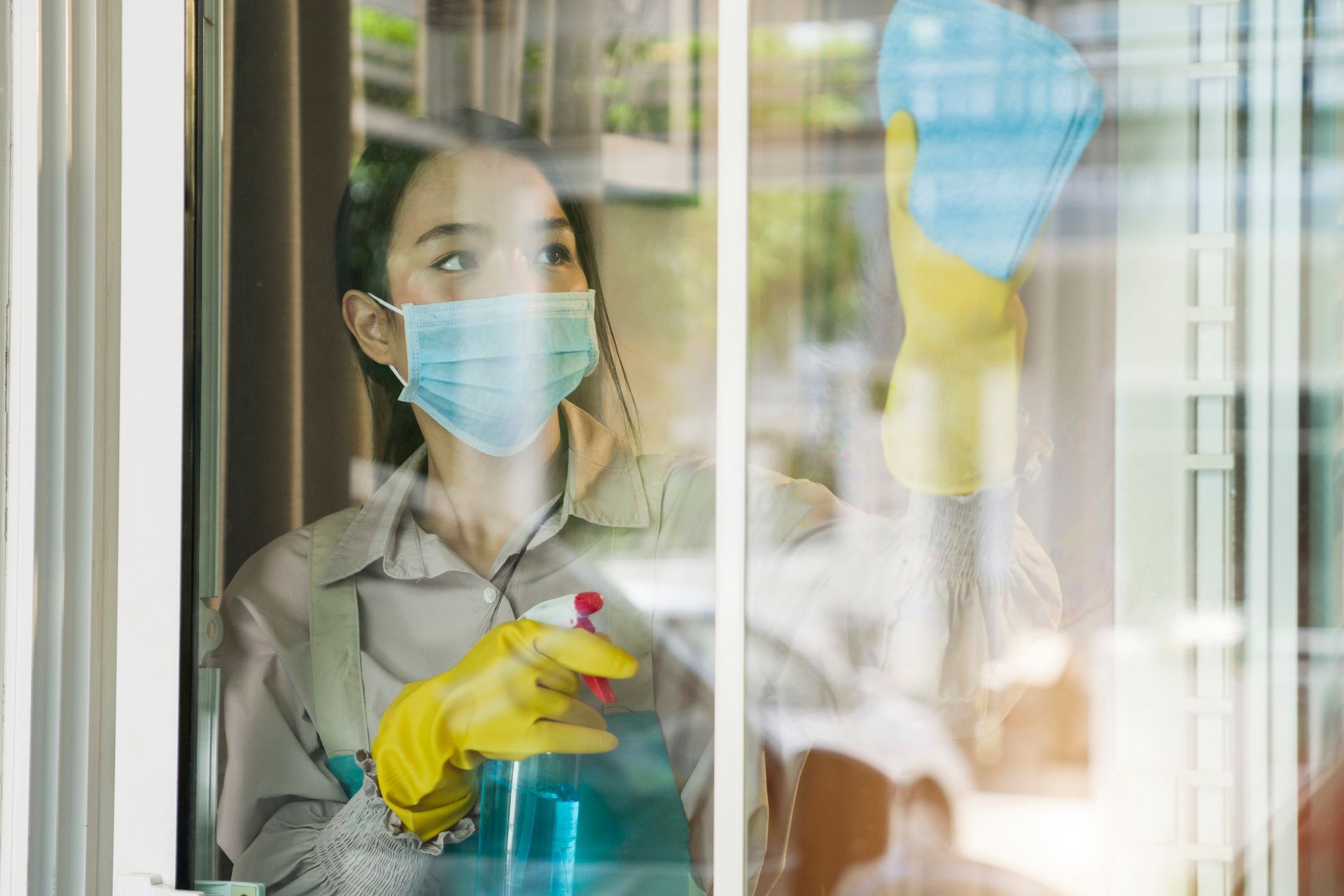 Masked cleaner spraying disinfectant by a window, wearing yellow gloves and holding a blue bottle.