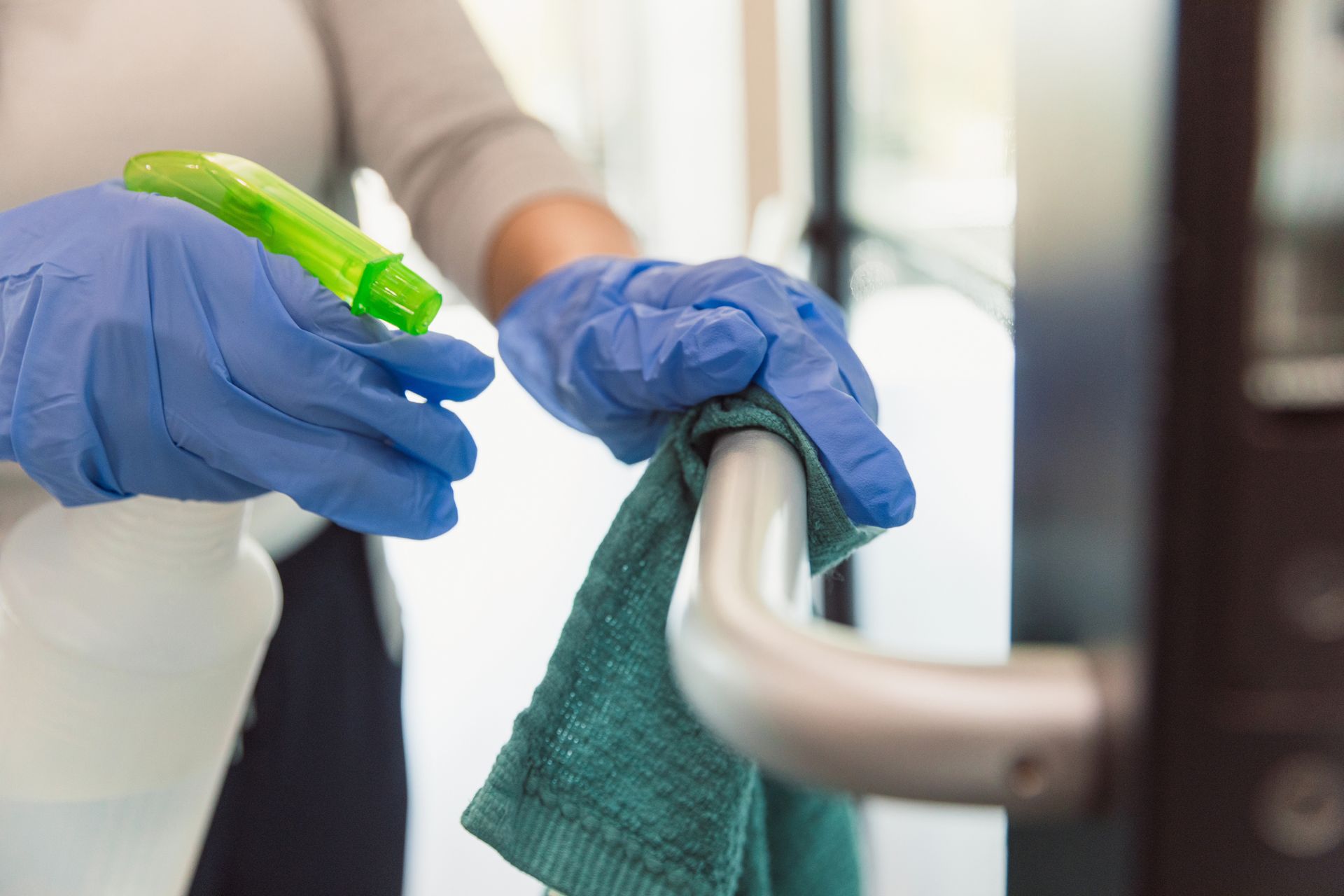 Gloved hands spraying cleaner and wiping a metal door handle with a green cloth