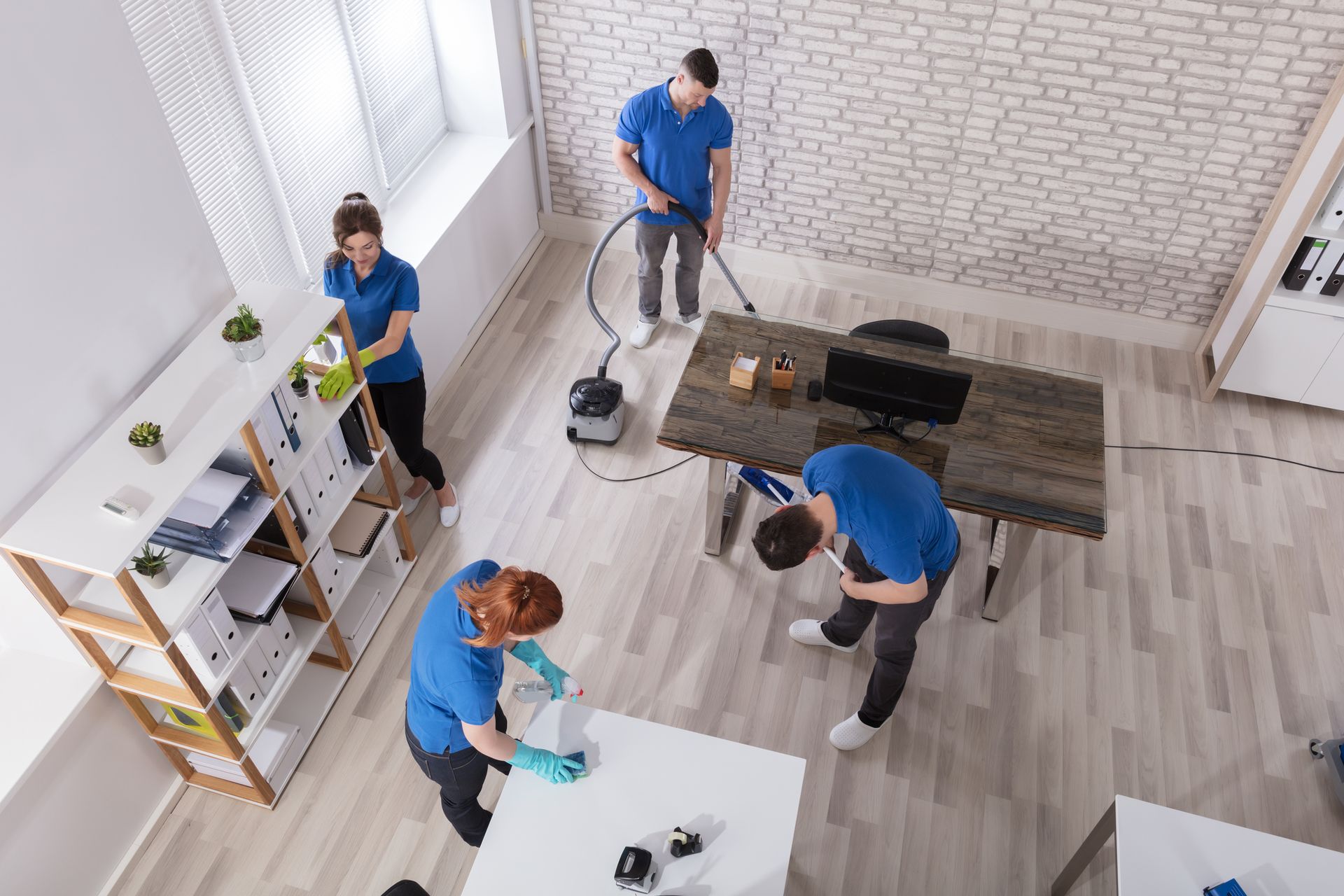 Three cleaners vacuum and wipe down a bright room with wood floors and a dining table.
