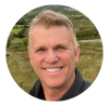 Smiling middle-aged man with short blonde hair, outdoors, wearing a dark shirt, in front of a grassy hillside.