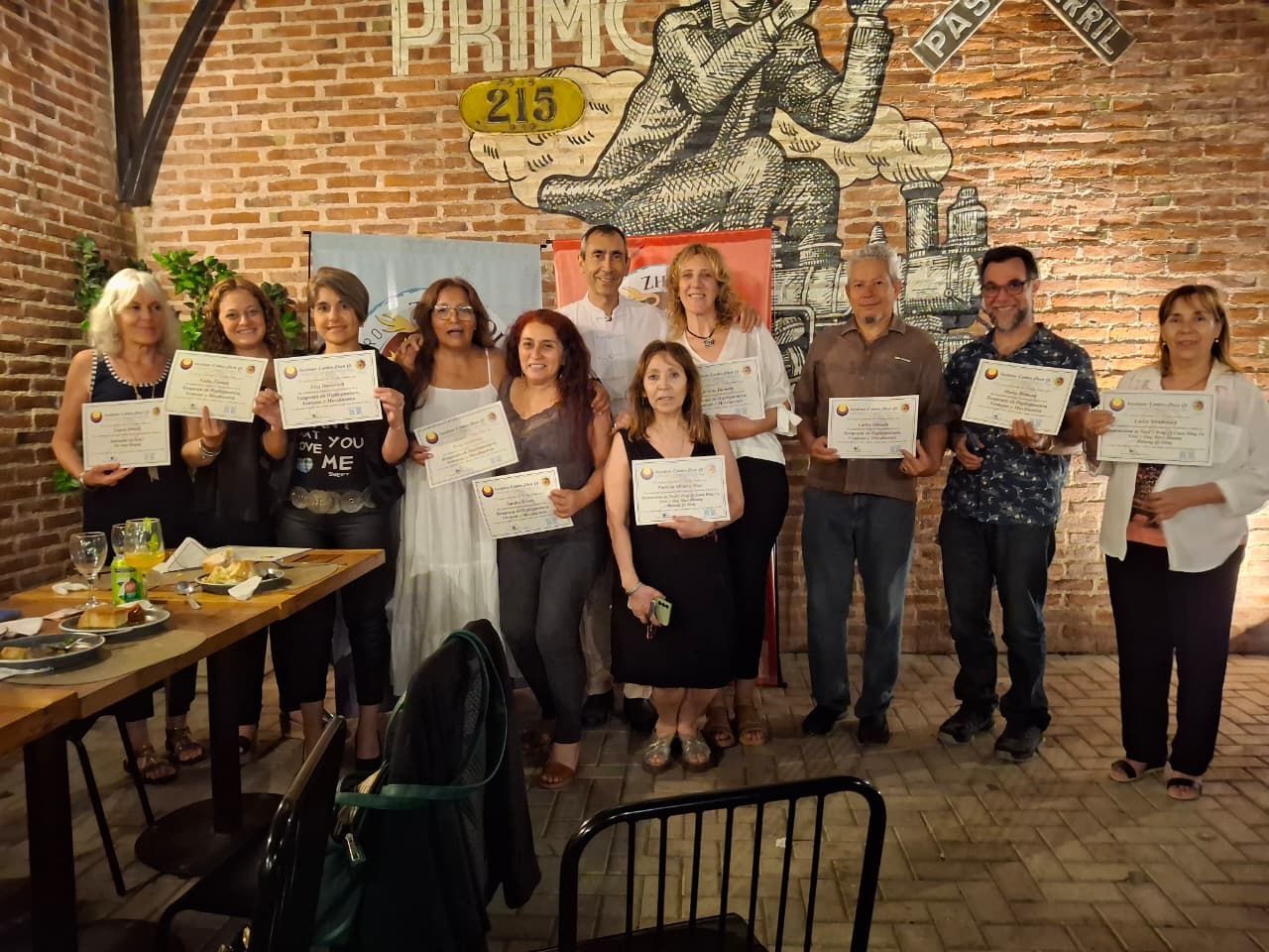 Grupo de personas sosteniendo certificados en una habitación con una pared de ladrillos y un mural.