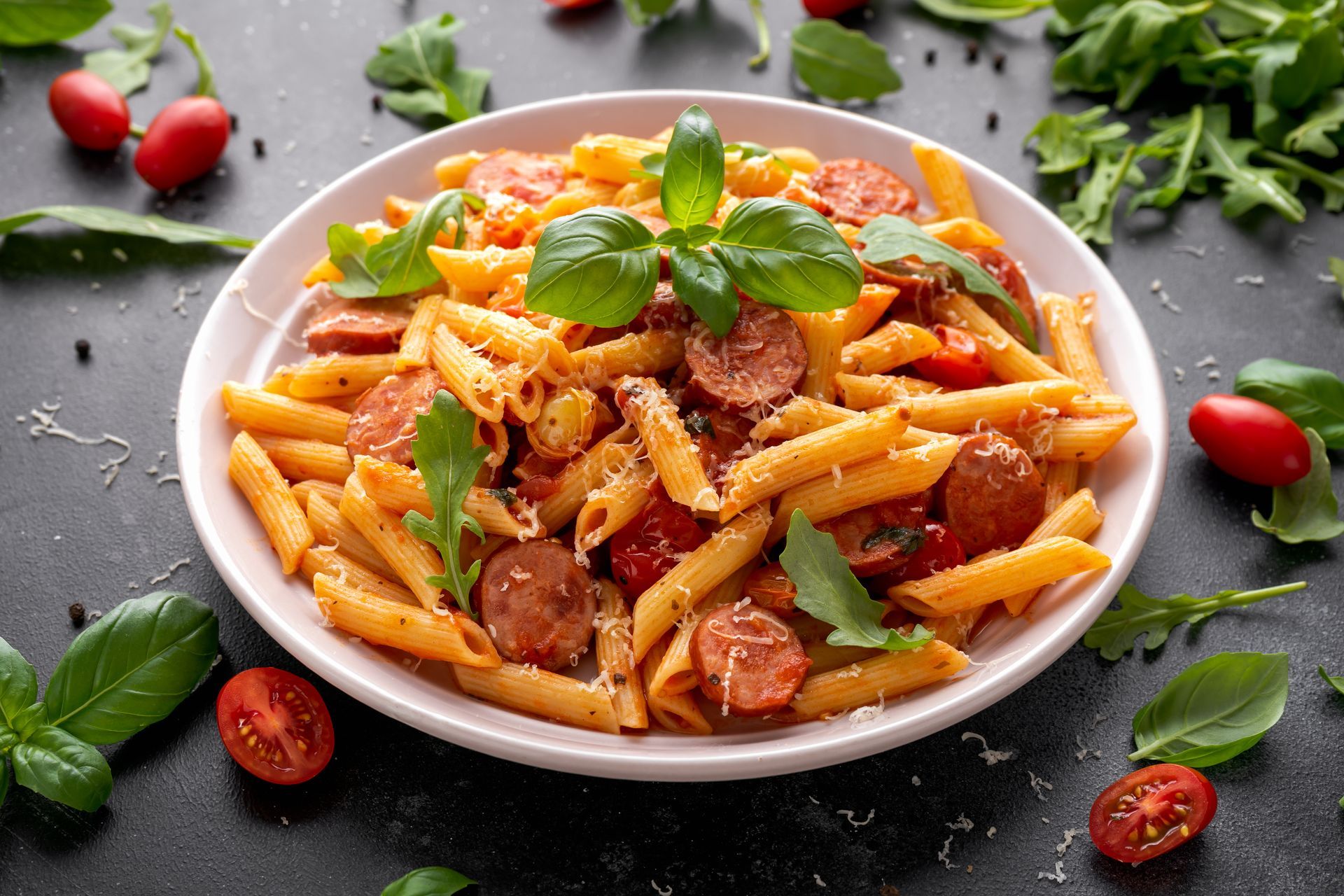 A plate of pasta with sausage , tomatoes and basil on a table.
